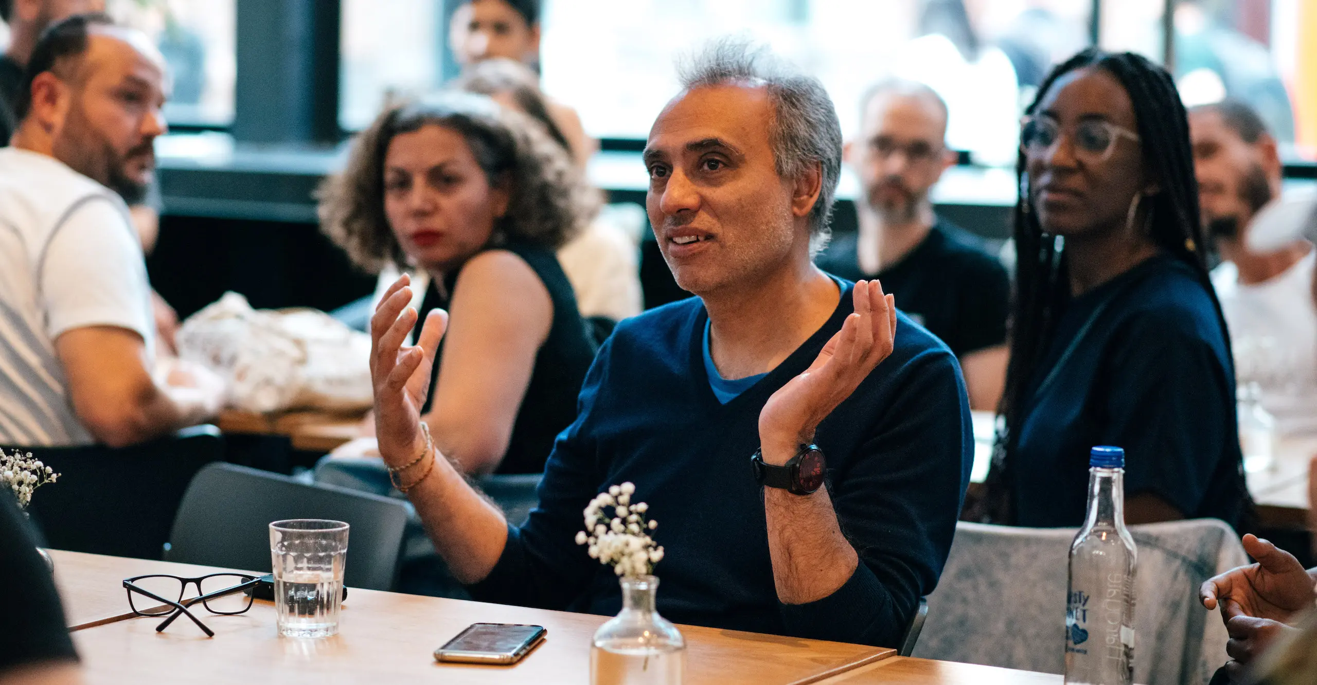 People sitting at cafe tables with one person facing the camera, hands gesturing as they speak.